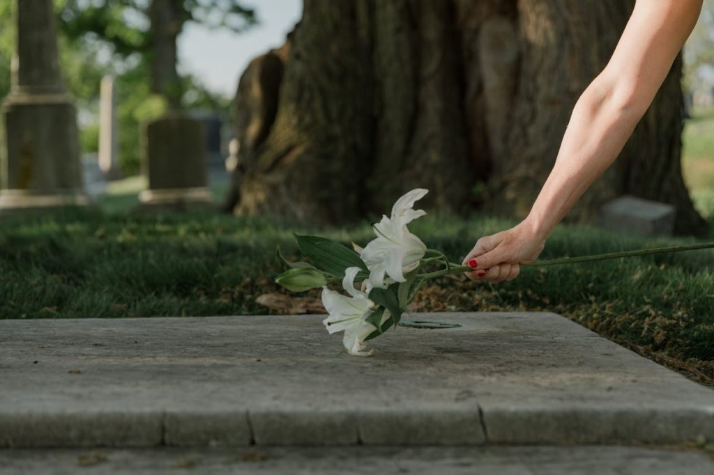 A person places a bouquet of white lilies on a gravestone in a cemetery with trees and headstones in the background.