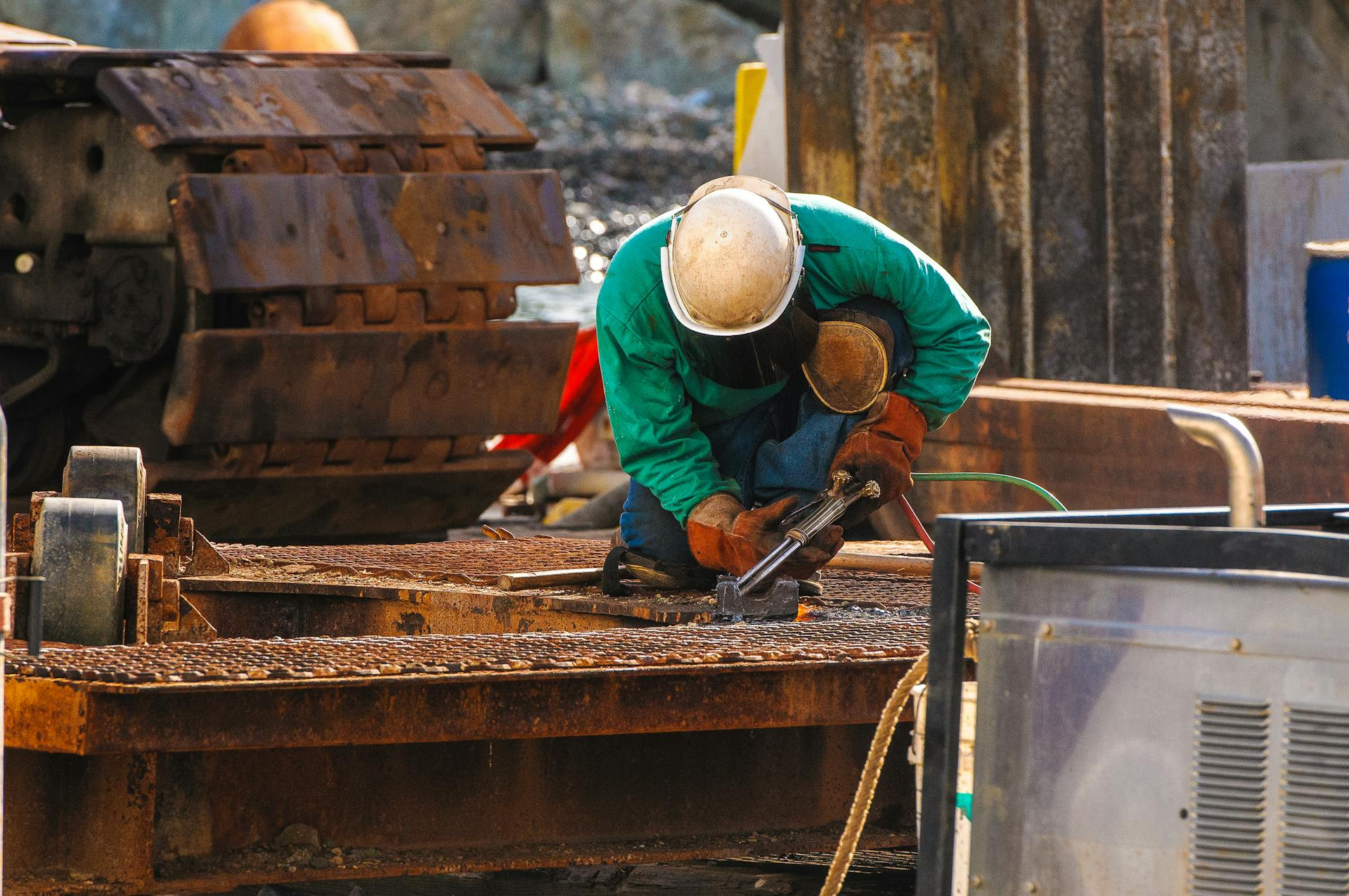 A worker in safety gear kneels on a metal platform while welding, surrounded by industrial equipment and machinery.