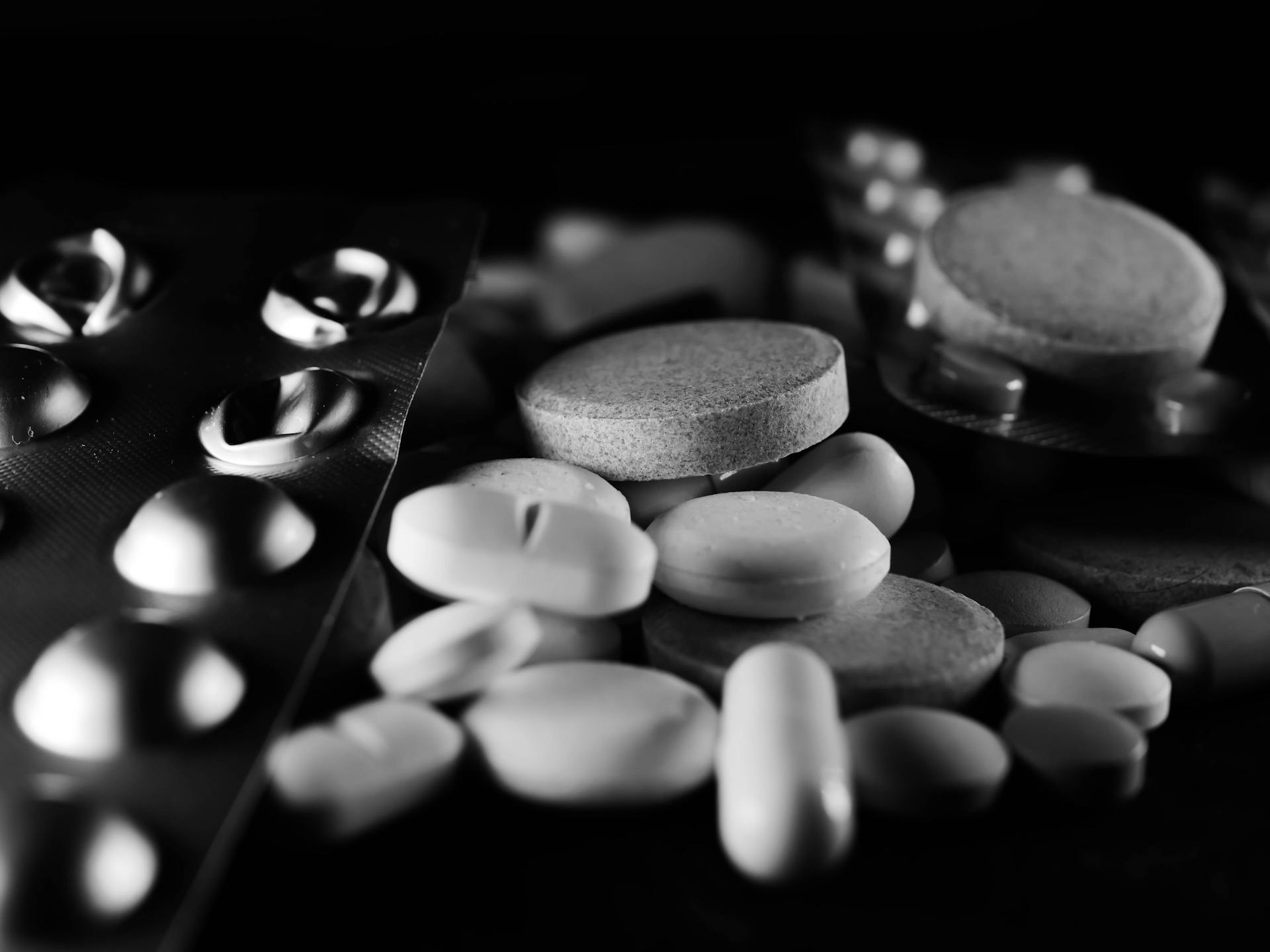 A variety of pills and tablets, some in blister packs and others loose, are displayed on a dark surface in black and white.