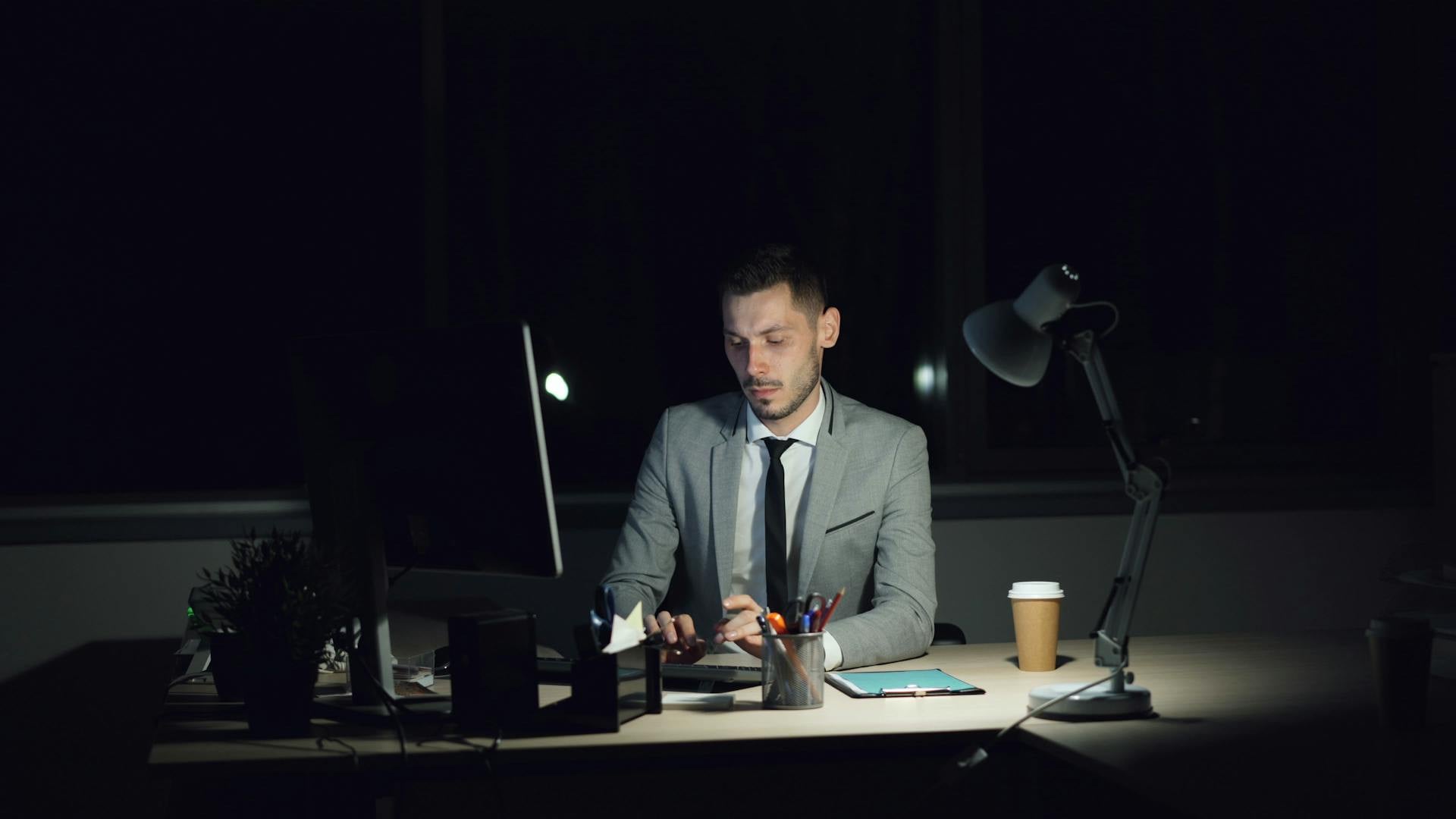 A man in a suit works at a desk in a dimly lit office at night, using a computer. A lamp, coffee cup, and office supplies are on the desk.