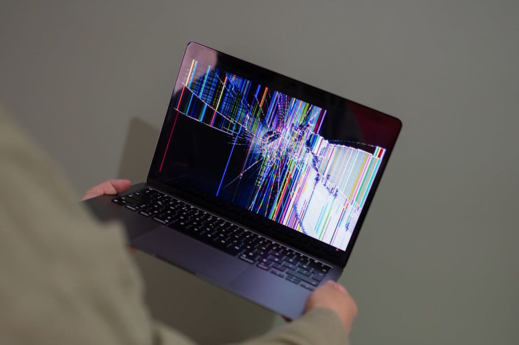 Person holding a laptop with a severely cracked screen displaying colorful vertical lines and distorted graphics.