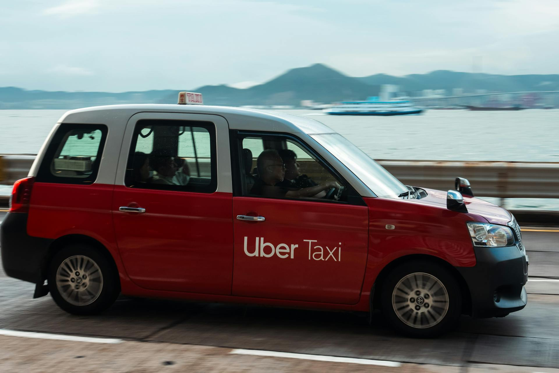 A red and silver taxi with "Uber Taxi" branding drives along a waterfront road with mountains and water visible in the background.