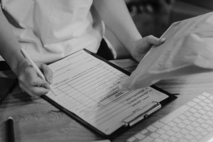 A person fills out a form on a clipboard while holding a large envelope, next to a computer keyboard on a wooden desk.