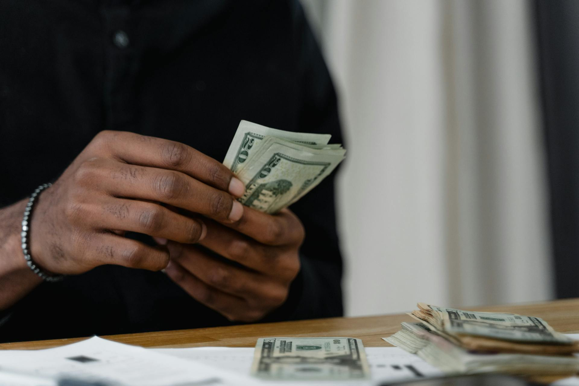 A person in a black shirt counts a stack of U.S. dollar bills at a wooden table with more cash and paperwork in the foreground.