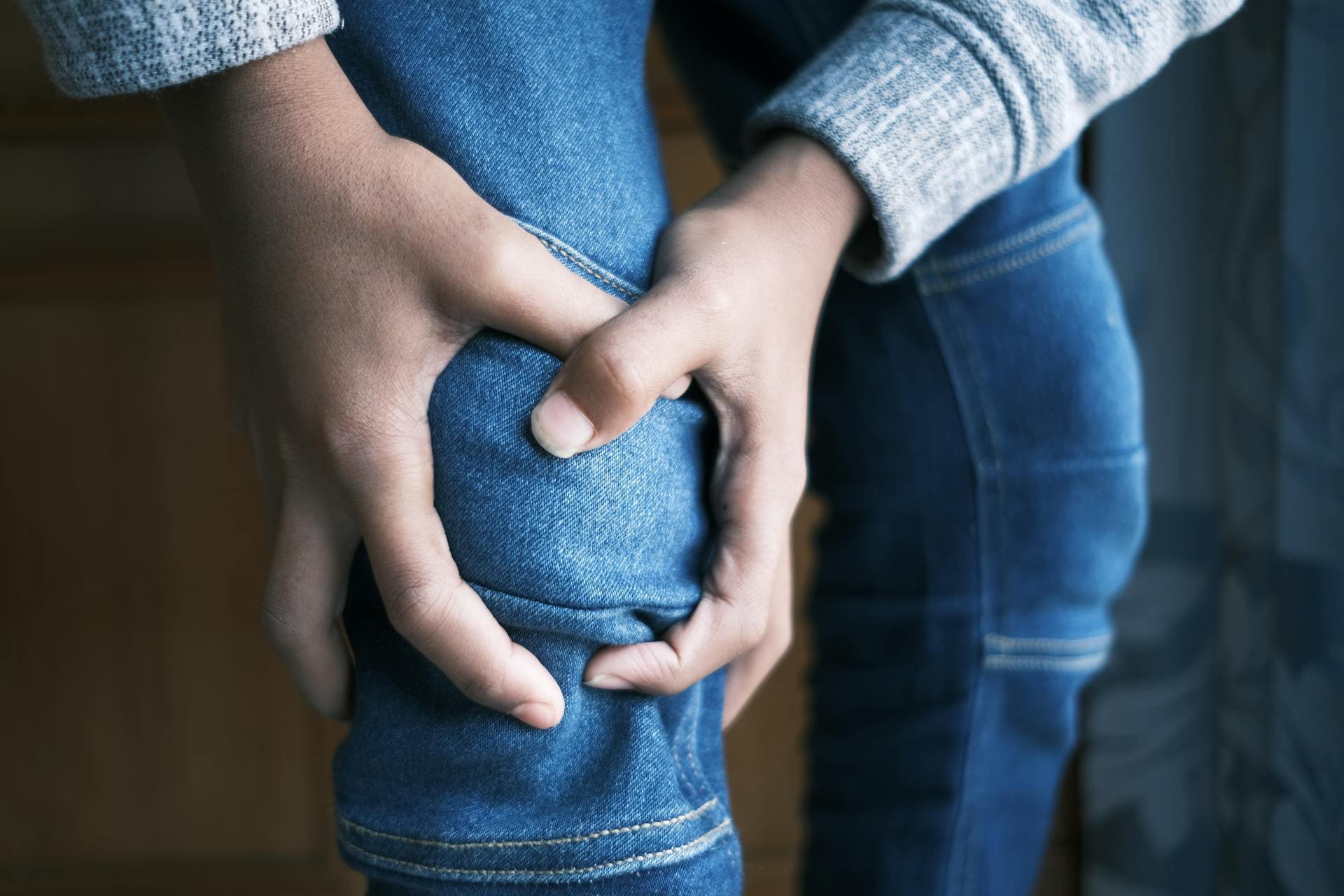 A person wearing blue jeans and a gray sweater holds their knee with both hands, suggesting discomfort or pain.