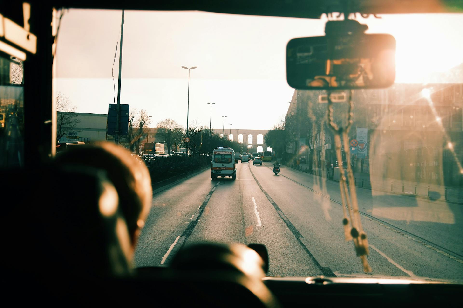View from inside a vehicle looking out at a sunlit road with cars, streetlights, and an aqueduct visible in the distance.