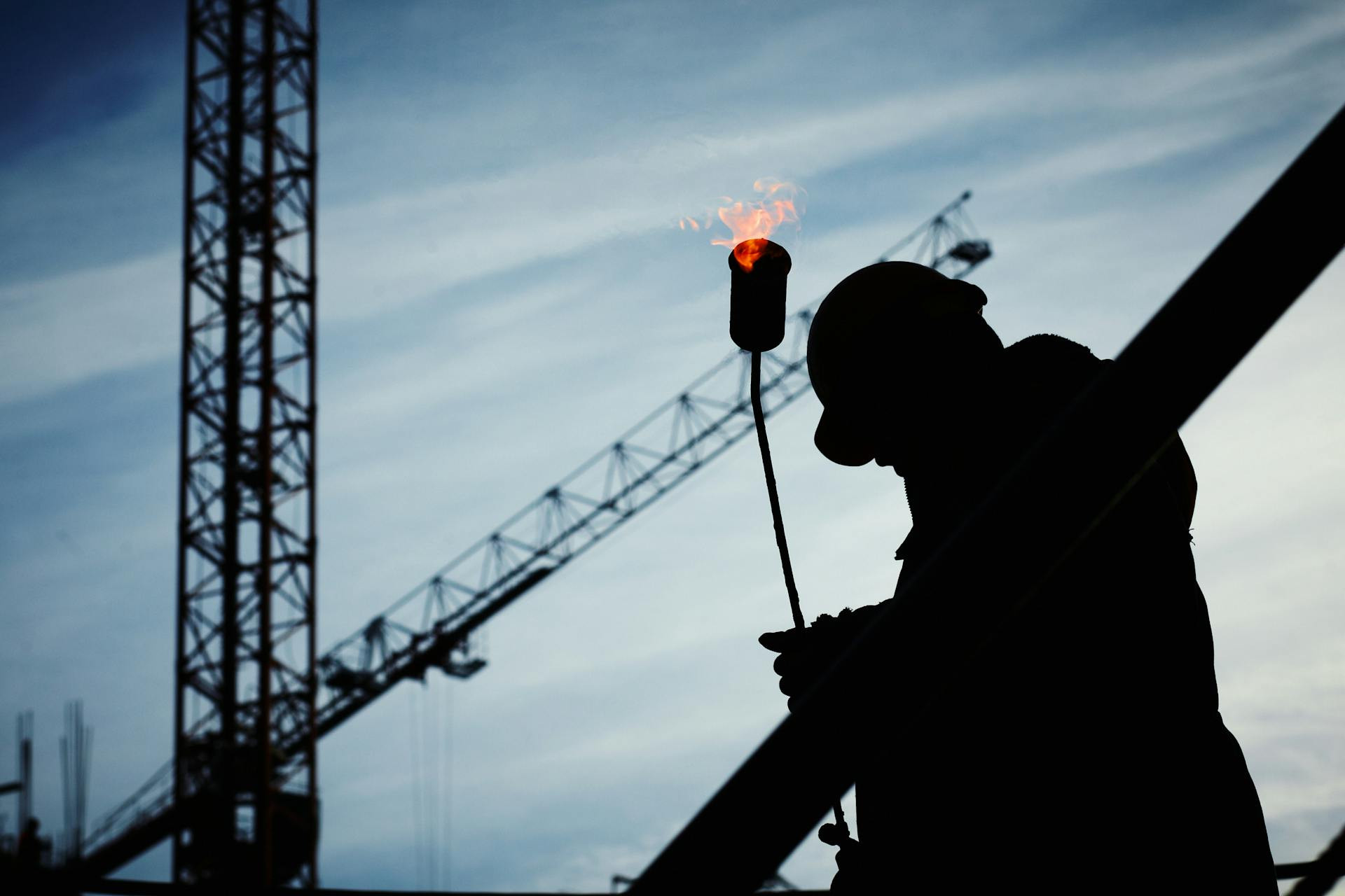 Silhouette of a construction worker holding a lit torch, with a crane and cloudy sky in the background.