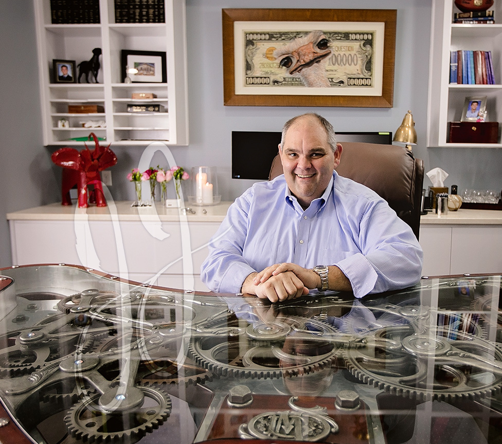 A man sits at a glass desk with visible gears underneath, in an office with shelves, books, flowers, and a framed artwork of a banknote on the wall behind him.