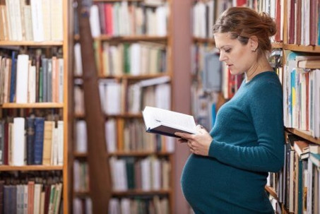 A pregnant woman stands in a library aisle, reading an open book while surrounded by bookshelves.