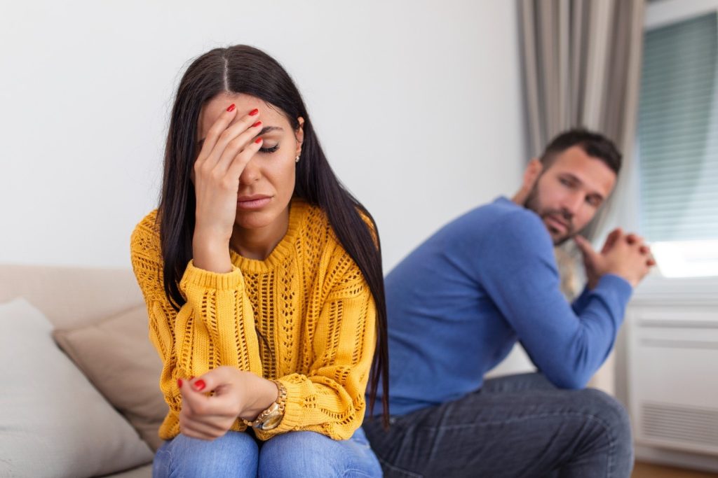 A woman sits with her hand on her face looking distressed, while a man sits behind her on the couch, looking away with a serious expression.