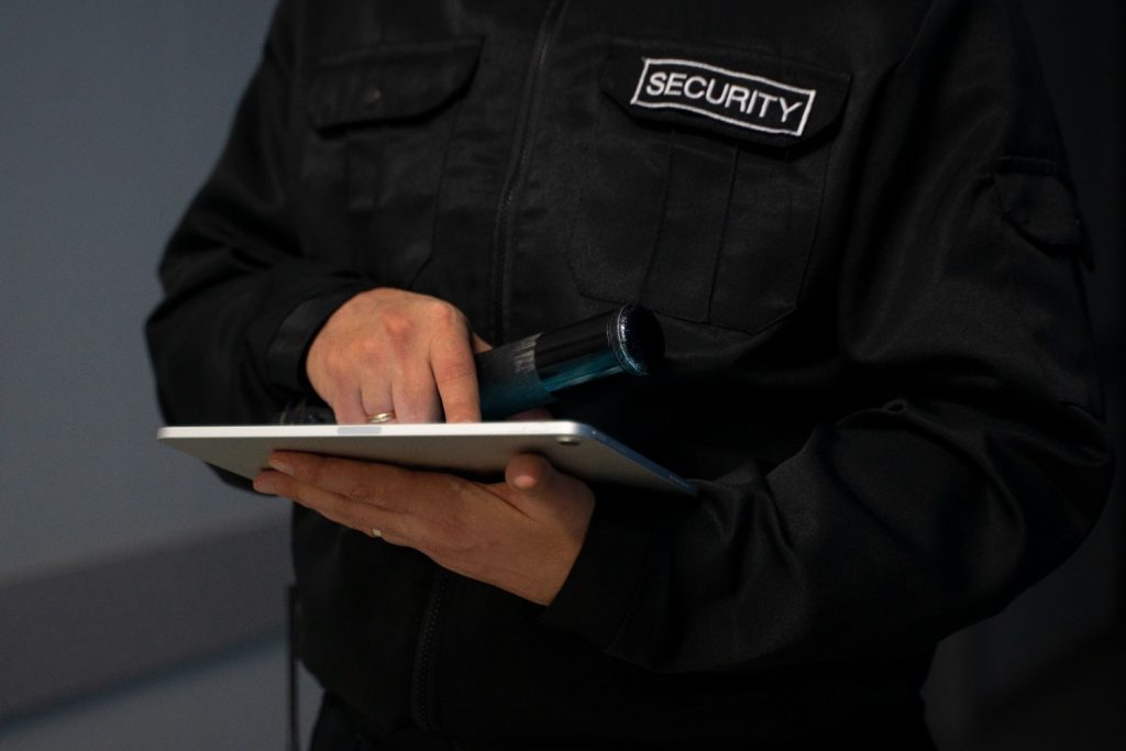 A person in a black security uniform holds a flashlight and uses a tablet.