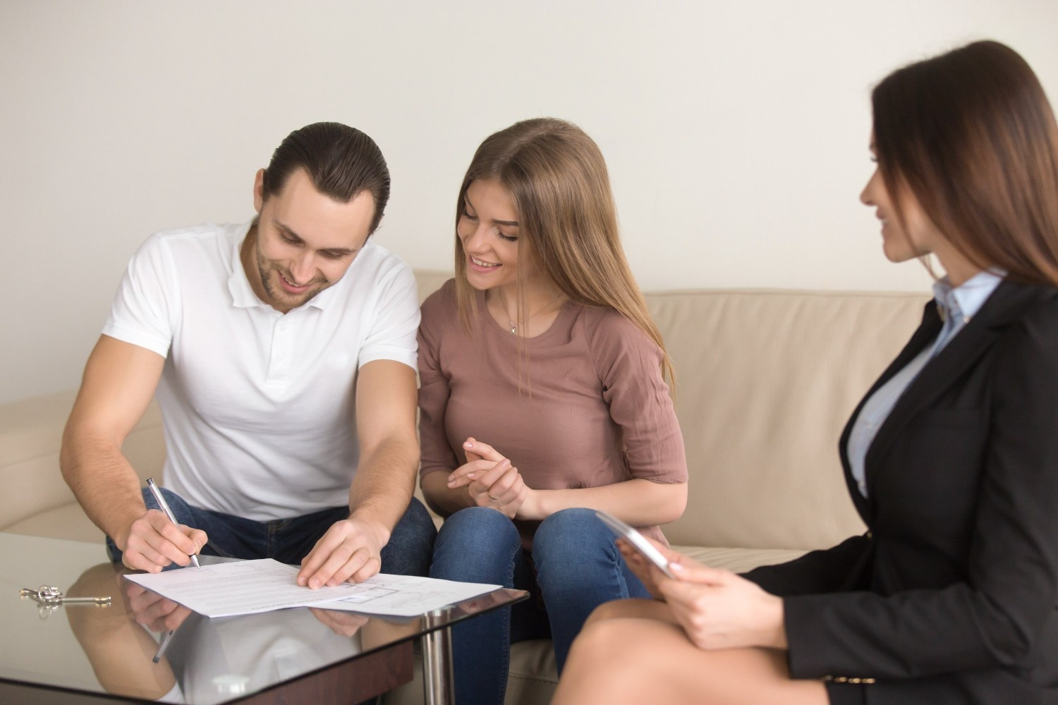 A man signs a document at a glass table while a woman sits beside him and another woman in business attire observes.