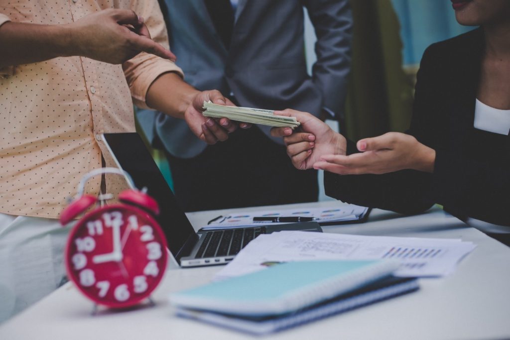 Three people in business attire exchange a stack of cash over a desk with documents, a laptop, and a red alarm clock.