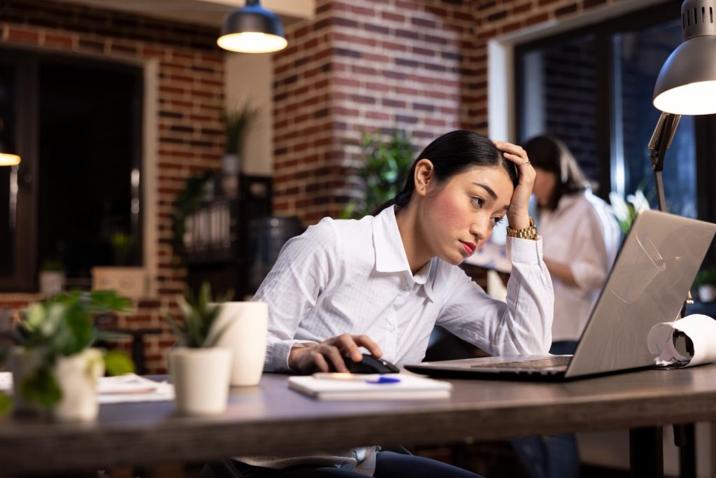 A woman sits at a desk, looking stressed while working on a laptop in a modern office with another person in the background.