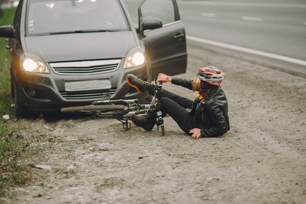 A person wearing a helmet lies on the ground next to a bicycle in front of a car with an open door on the side of a road.