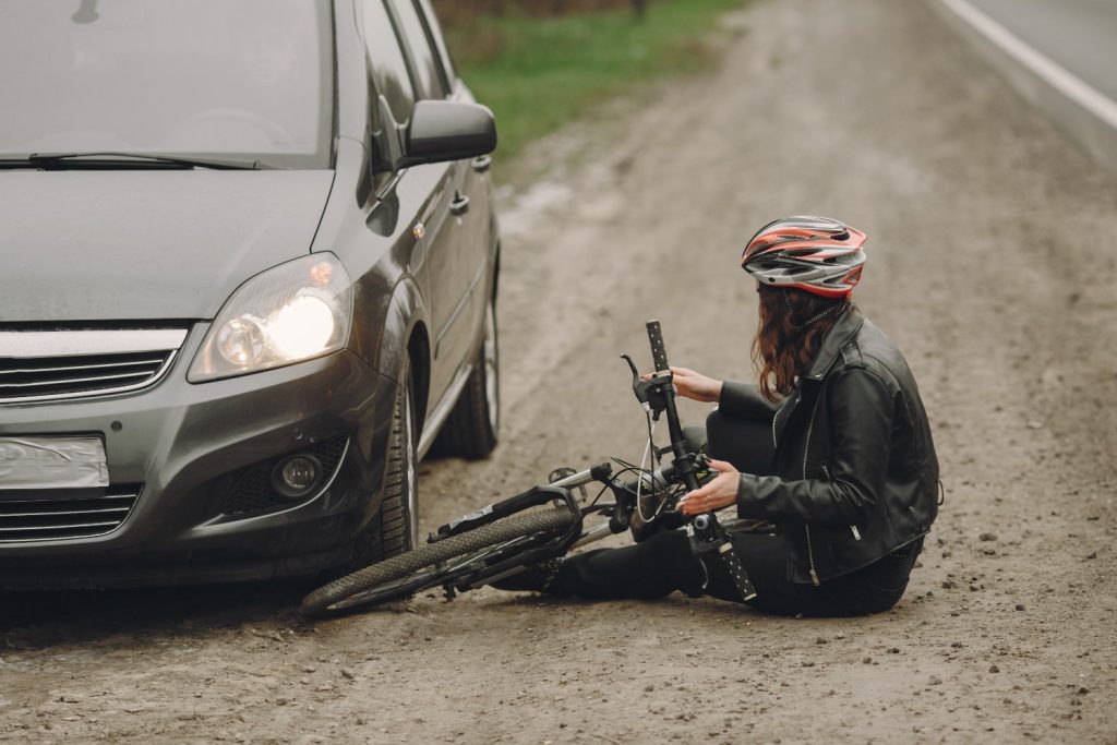 A person wearing a helmet sits on the ground next to a fallen bicycle and a car by the roadside, suggesting a possible accident.