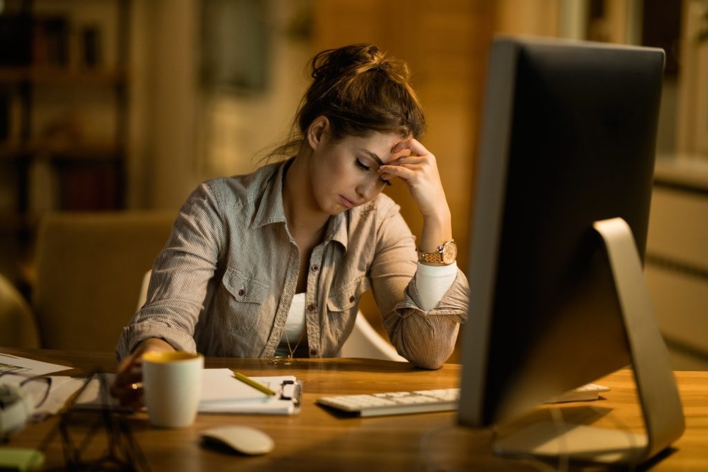 A woman sits at a desk in front of a computer, looking tired and holding her head with one hand, with a notebook, pen, and coffee mug nearby.