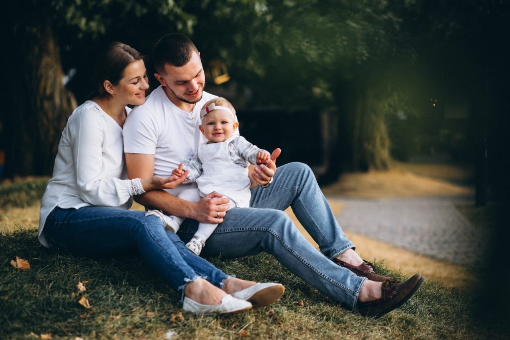 A man, woman, and baby sit together on a grassy slope outdoors, all smiling and wearing casual white and denim clothing.