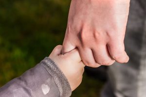 An adult hand holding the thumb of a child's hand, both wearing long-sleeved clothing, with a blurred green background.